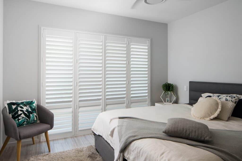 White wooden shutters in bedroom with grey chair and grey throw blanket